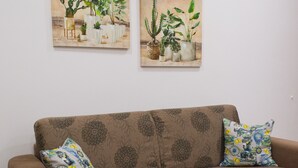 Un divano marrone con motivi floreali, due cuscini decorati e due quadri con piante appese al muro.