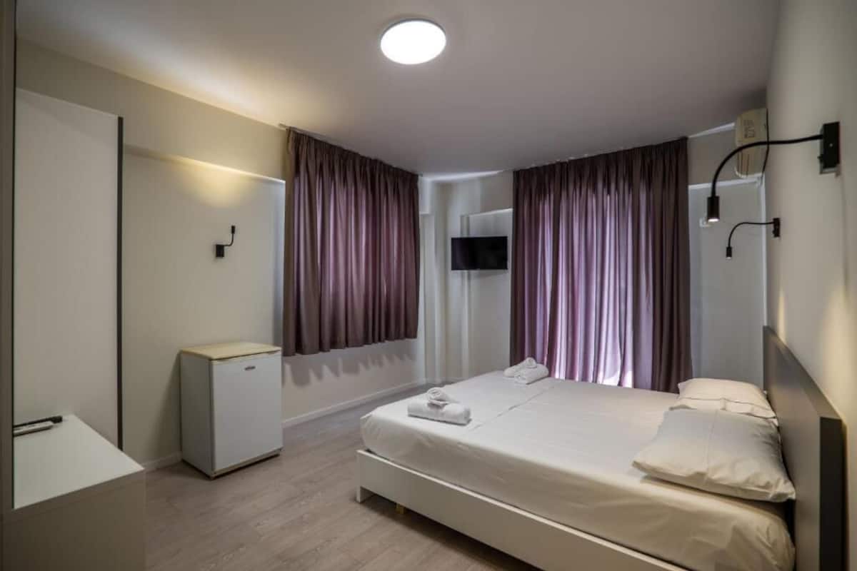 A modern hotel room with a single bed, a small desk, a television, and a minibar.