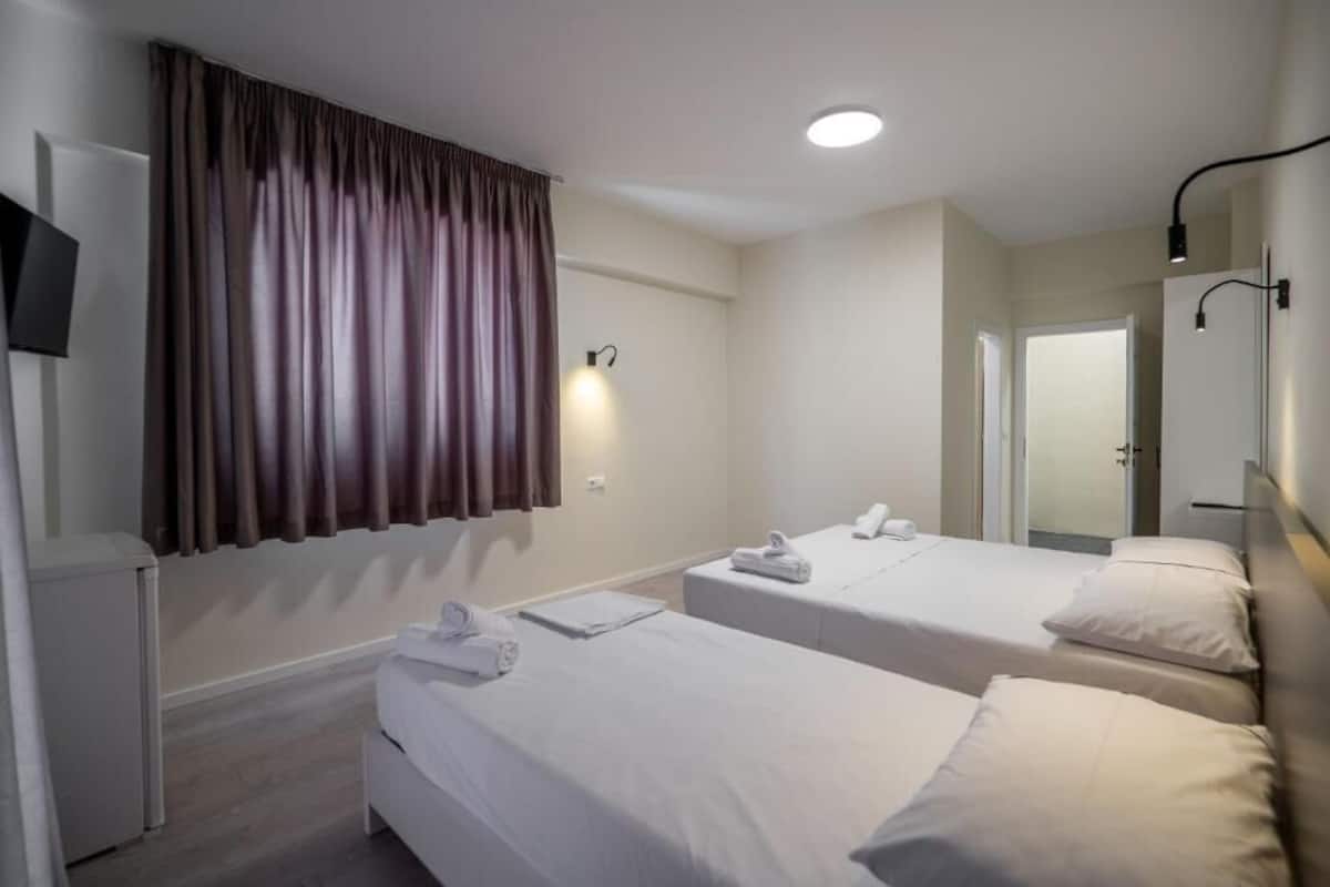 A hotel room with two single beds, a small bedside table with a lamp, a TV mounted on the wall, and a large window with curtains.
