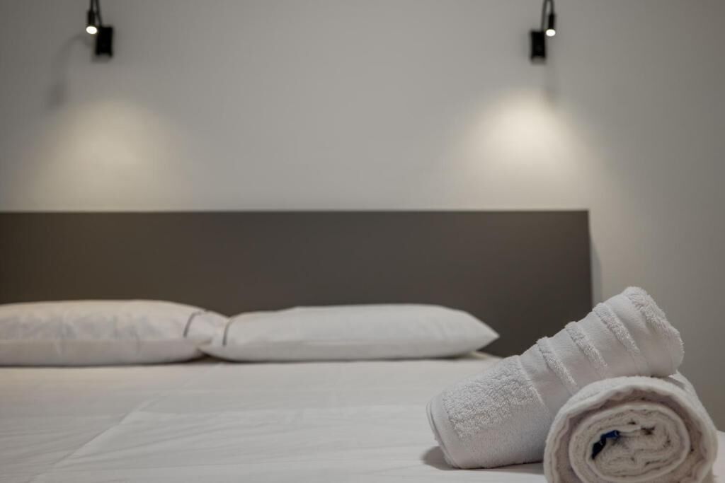 A neatly made bed with white linens and pillows, a rolled towel on the side, and two wall-mounted lights.