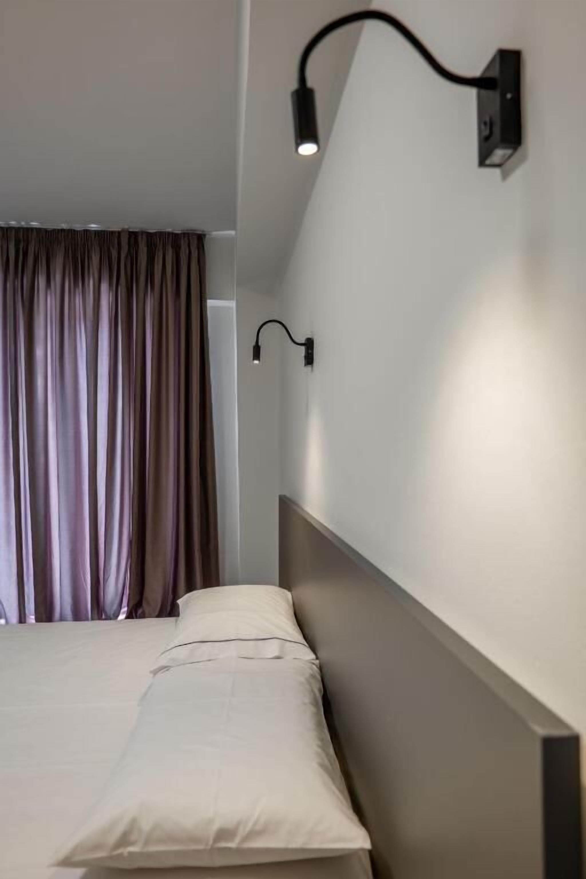 A hotel room with a bed, a wall-mounted light fixture, and a window with curtains.