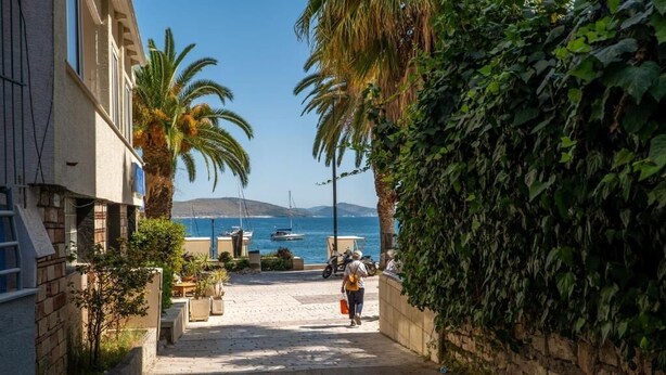A narrow street leading to a harbor with boats, palm trees, and a stone wall on one side. - Meraki Rooms (Sarande)