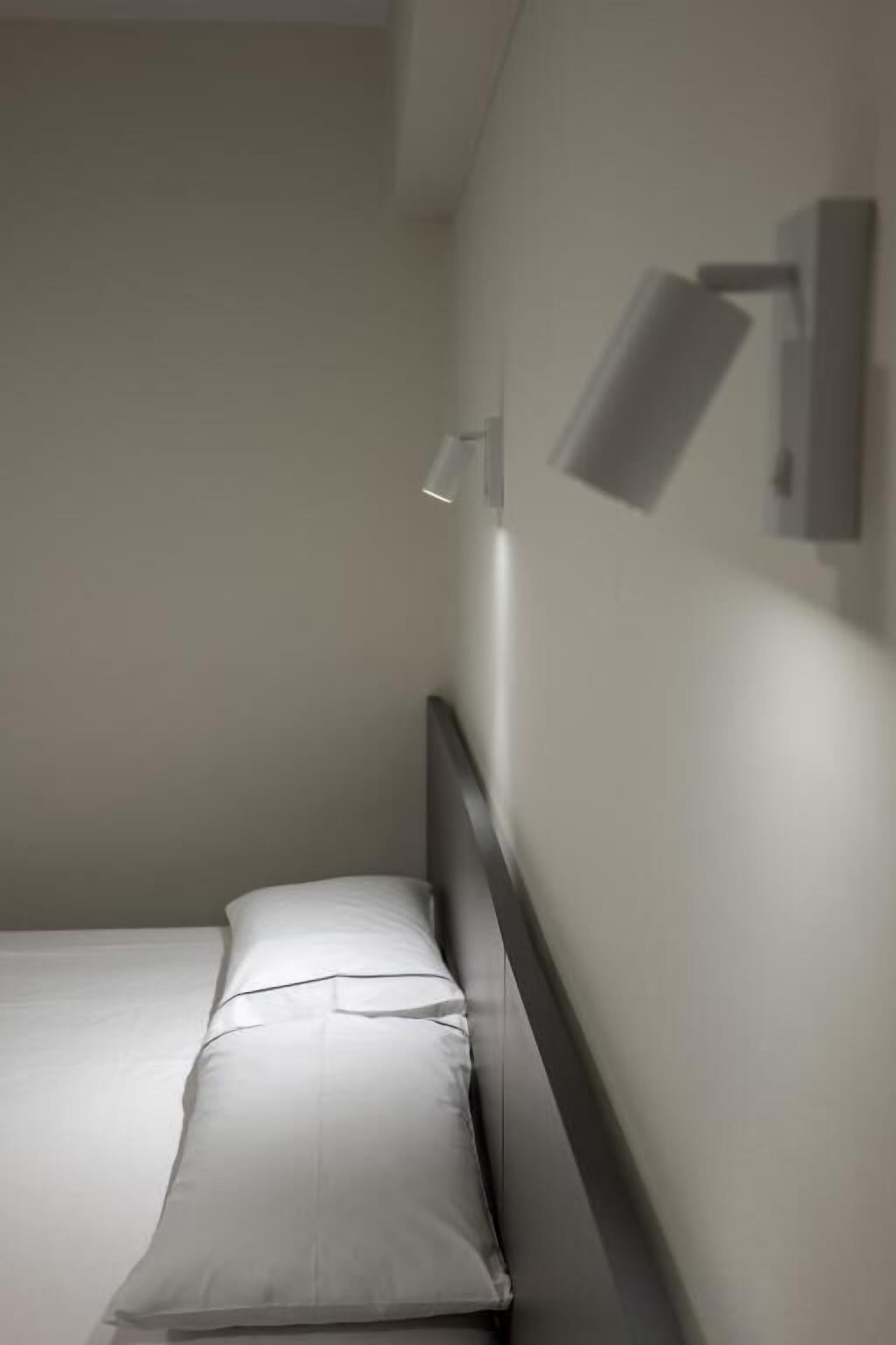 Et lille, aflangt værelse med en enkelt seng, et natbord og en lampe monteret på væggen.