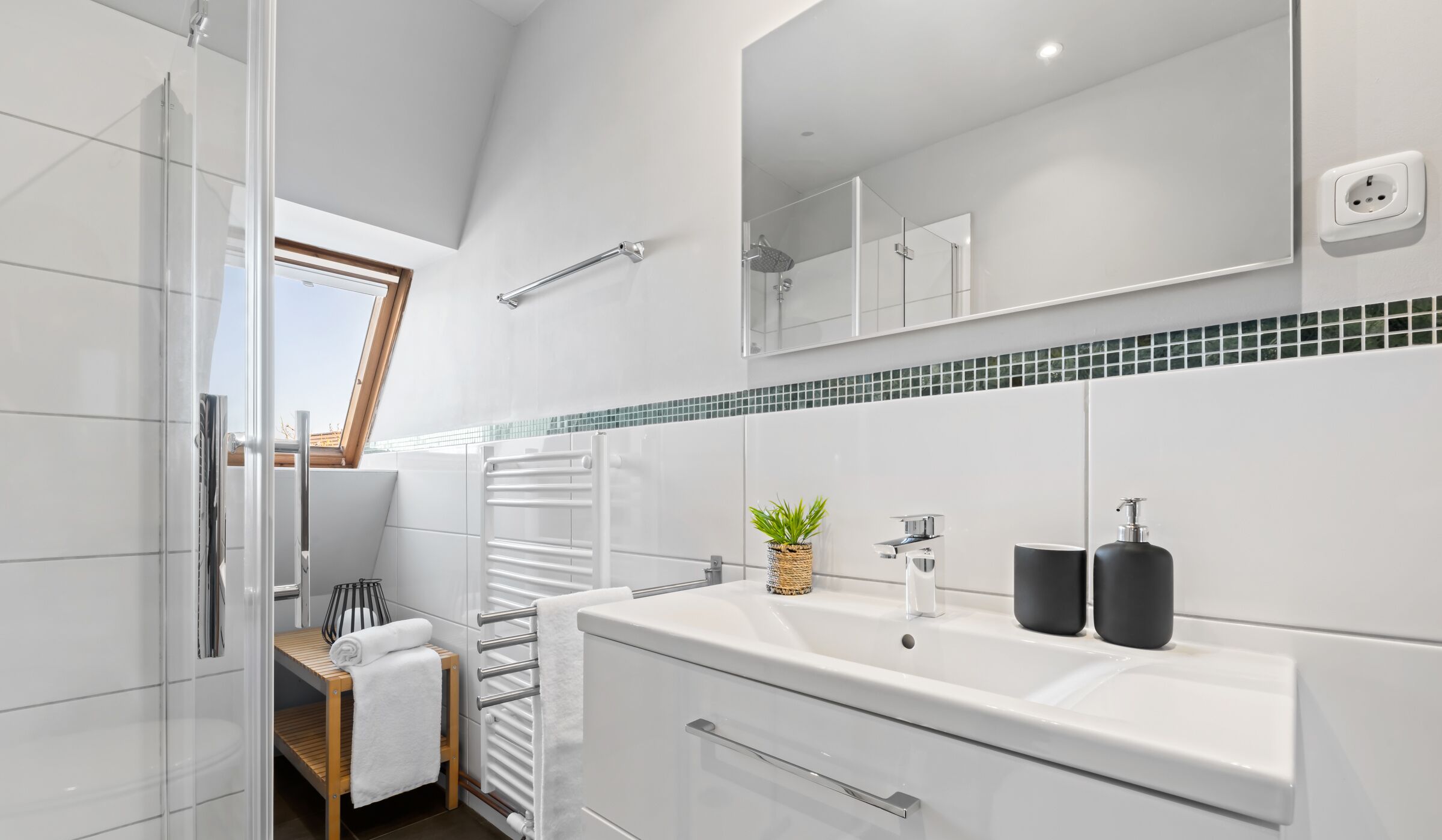 A modern bathroom with a glass shower enclosure, a white sink with black soap dispensers, a towel rack, and a small plant.
