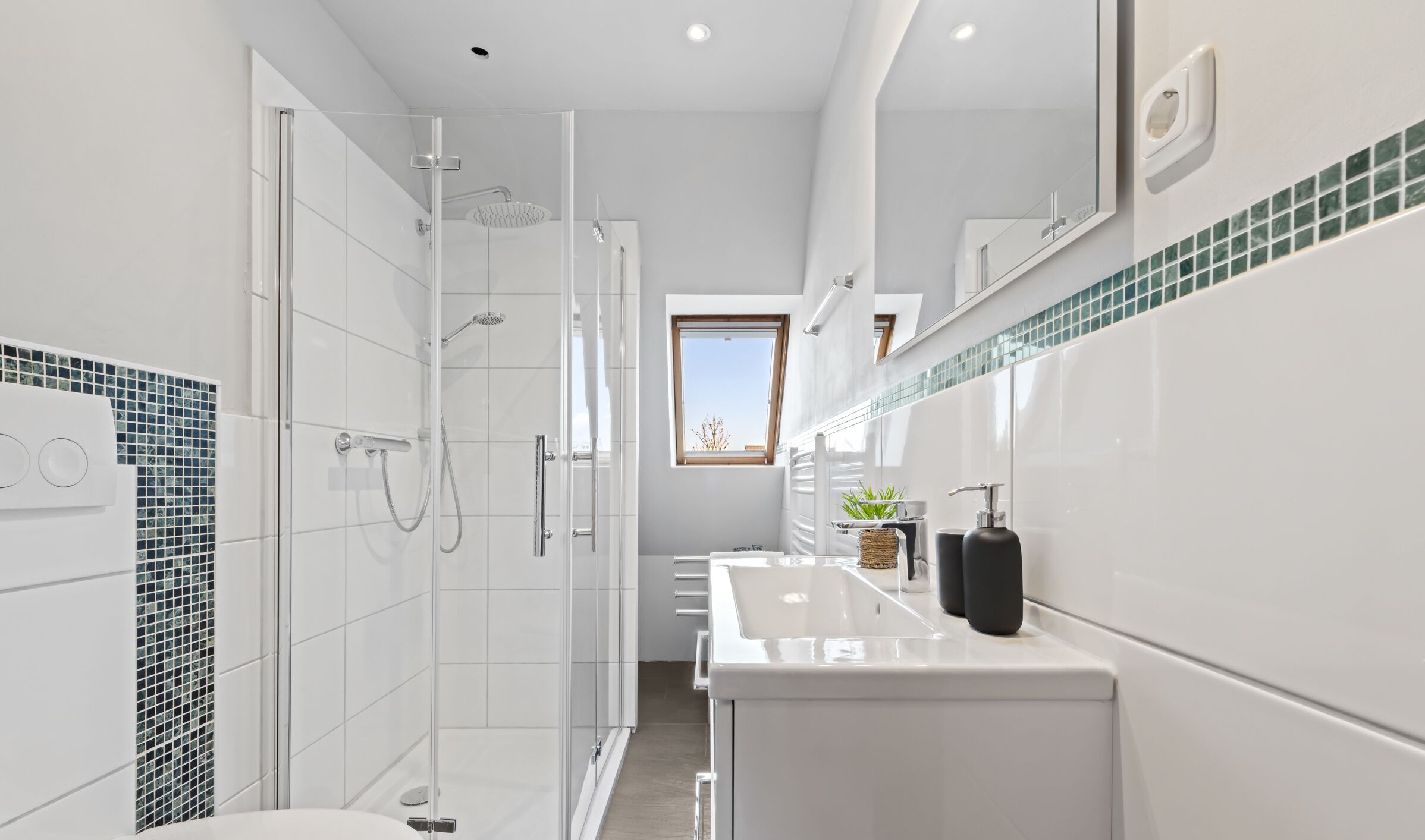 Ein modernes Badezimmer mit einer in Glas eingefassten Dusche, weißen Fliesen, einem großen Spiegel und einem Fenster mit Tageslicht.