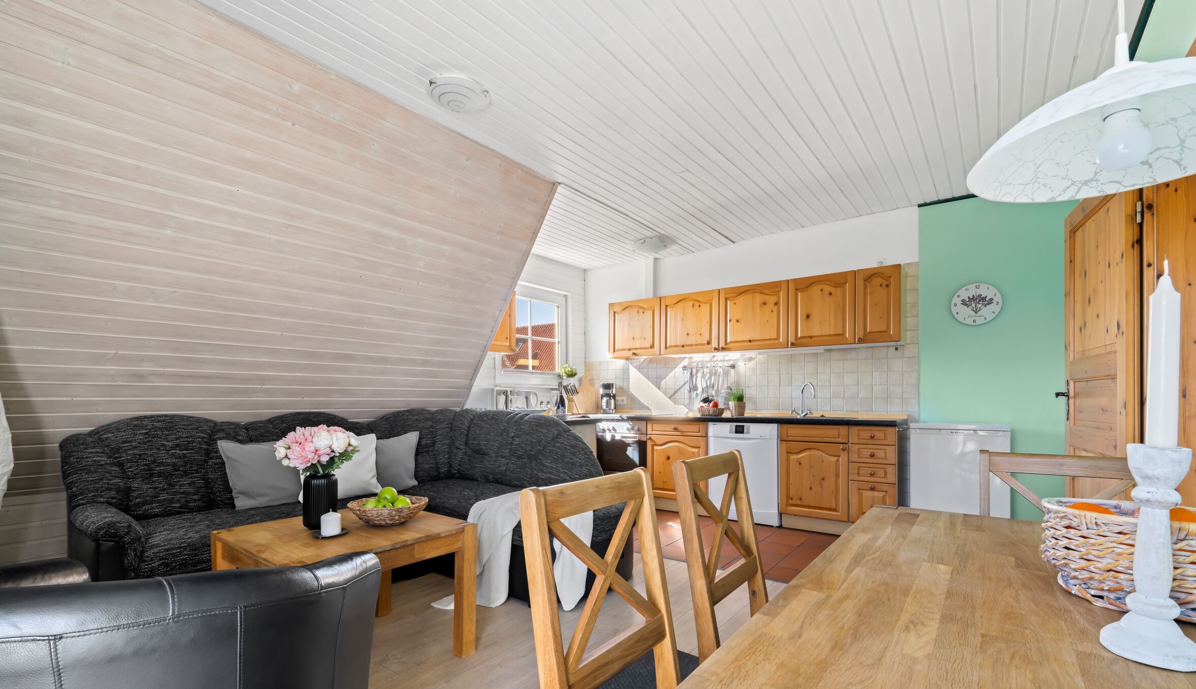 A cozy living room with a sofa, a wooden coffee table, and a dining table with chairs. A kitchen with wooden cabinets and a window is visible in the background.