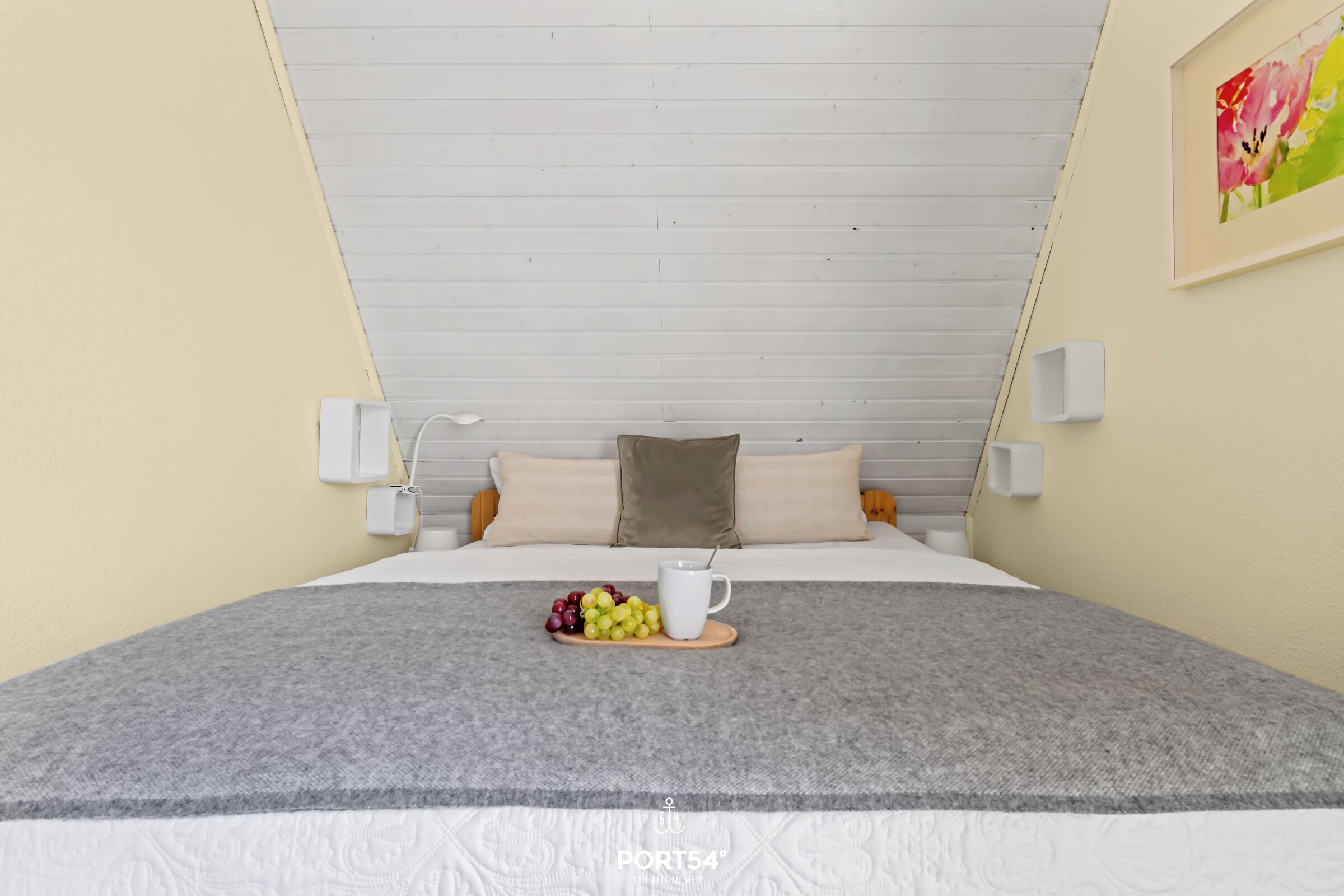 A modern bedroom with a sloped ceiling, a bed with a grey blanket, a wooden bedside table with a fruit bowl and a white mug, and a painting on the wall.