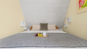 Une chambre moderne avec un plafond en pente, un lit recouvert d’une couverture grise, une table de chevet en bois sur laquelle se trouve un panier de fruits et une tasse blanche, et un tableau accroché au mur.