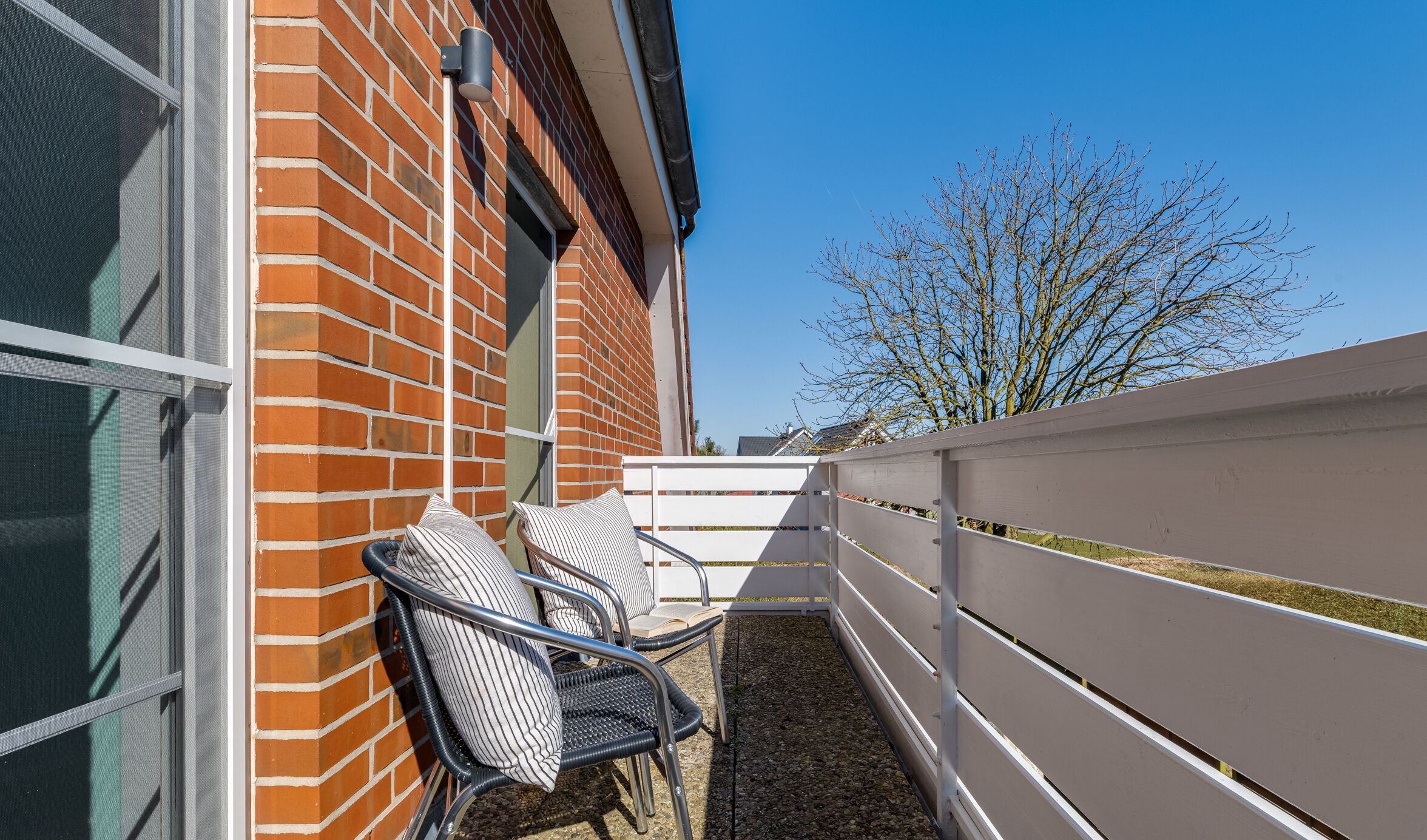 Ein Balkon mit zwei Korbstühlen, einem weißen Zaun und einem Backsteinhaus.