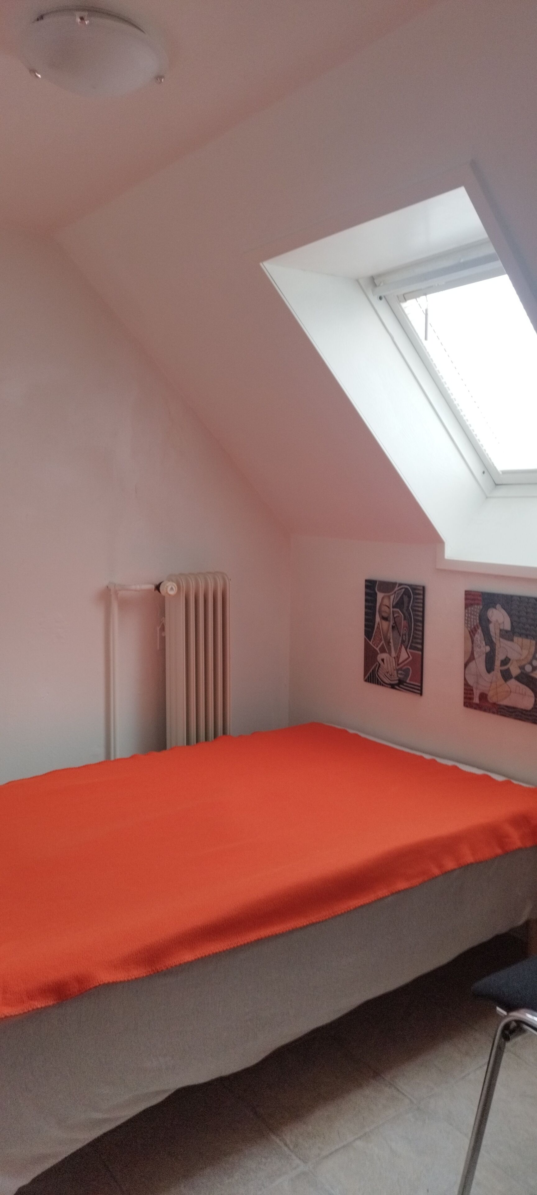 Een kleine eenpersoonskamer met een schuine plafond en dakraam, een radiator, en twee abstracte schilderijen aan de wanden.