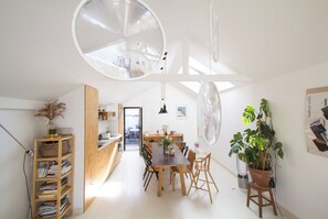A modern kitchen with a wooden dining table, chairs, and a large circular mirror. There is a bookshelf with books and a potted plant.
