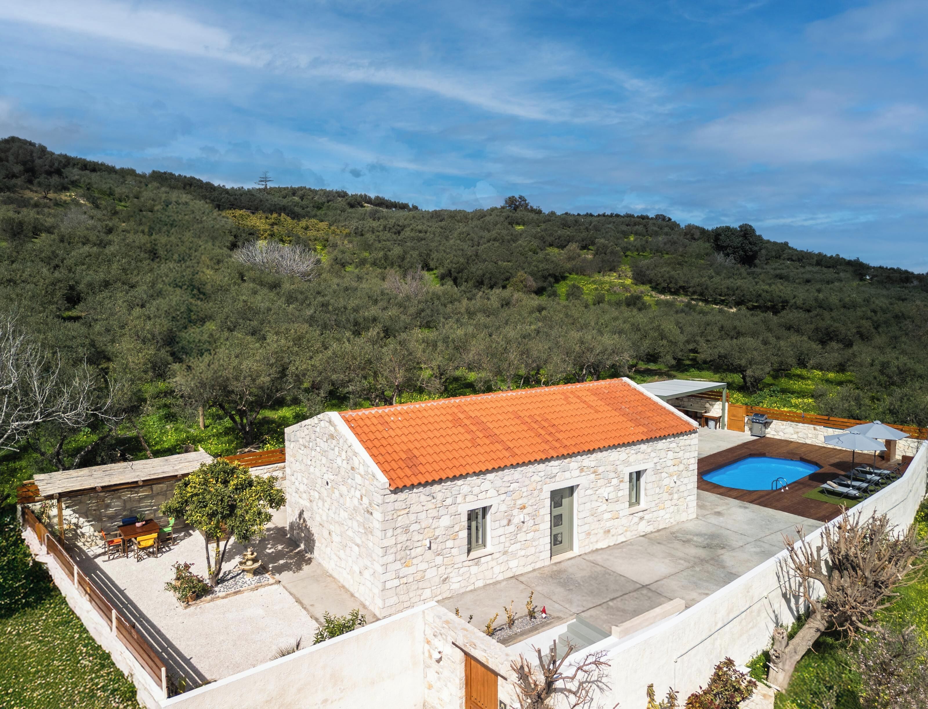 A stone building with a red-tiled roof, a swimming pool, and a view of a forested hillside.