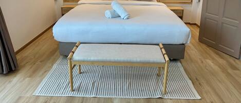 A neatly made bed with a white comforter and two pillows, a wooden bench at the foot of the bed, a woven headboard, and a wall with circular woven wall hangings.