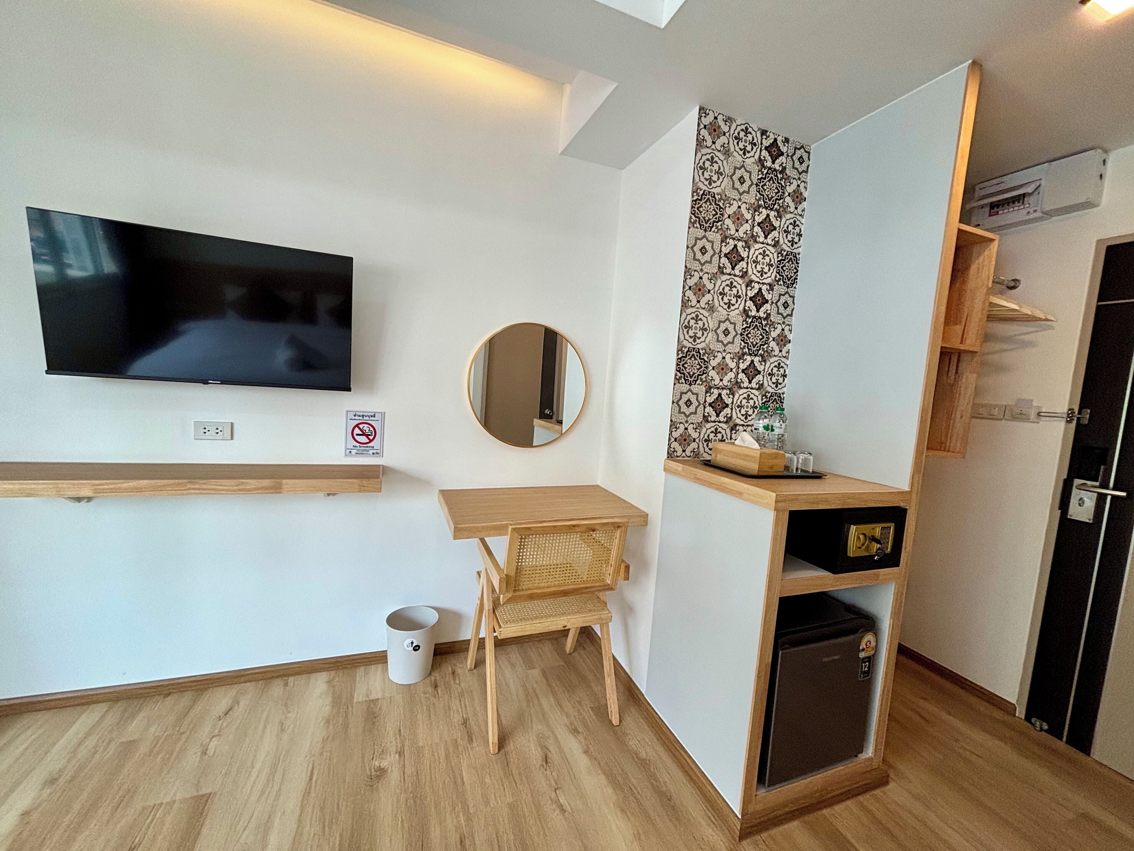 Ein modernes Zimmer mit Flachbildfernseher, runder Spiegel, kleinem Holztisch, Korbstuhl und integriertem Minikühlschrank.
