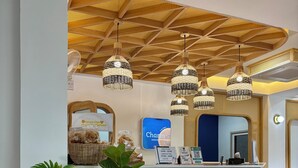 Een hotel receptie met een houten plafond, hangende rieten kroonluchters, een balie met een bordje met de tekst 'Charm Hostel' en grote potplanten.