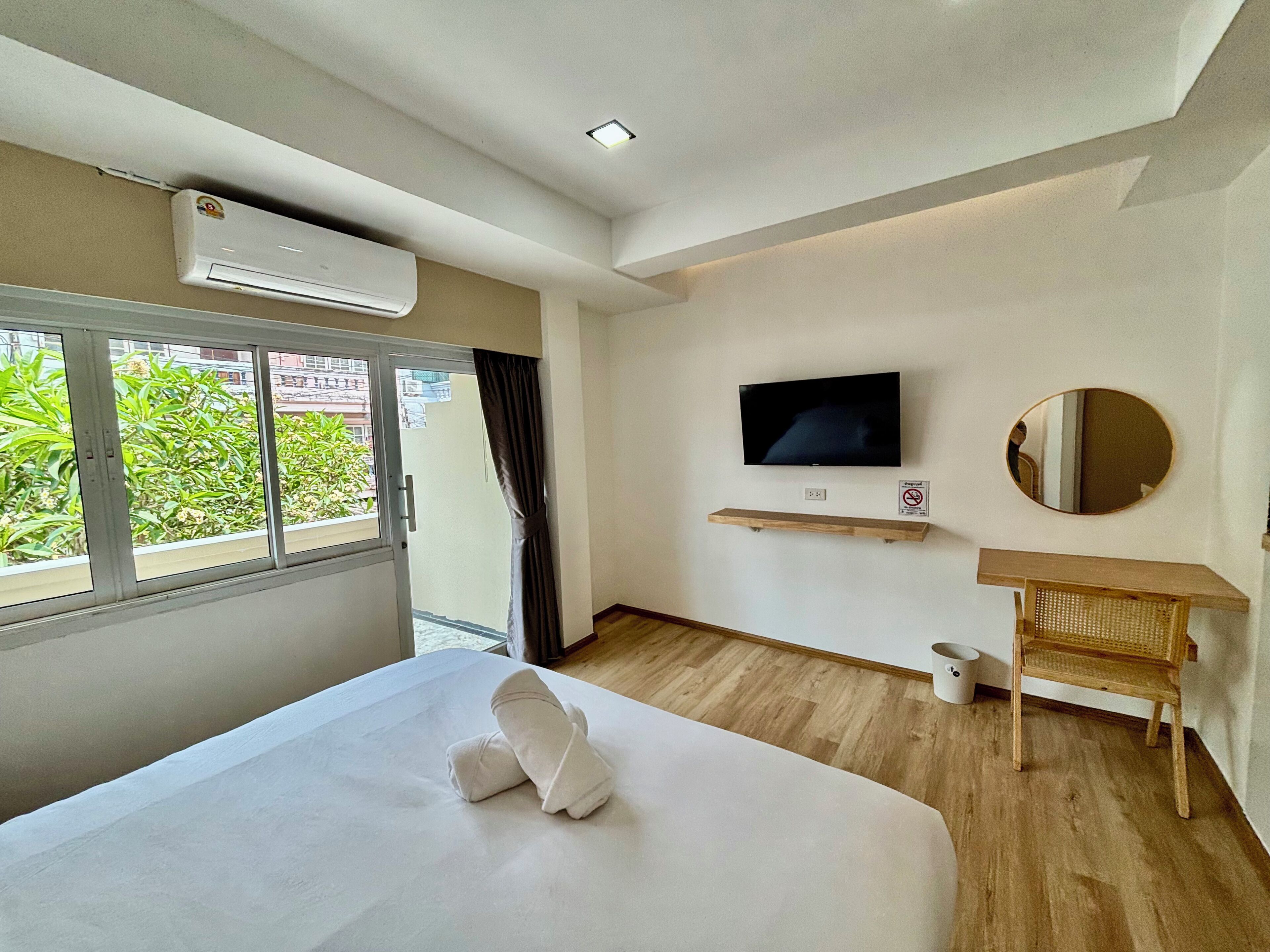 A hotel room with a bed, a TV, a mirror, a wooden shelf, and an air conditioning unit.