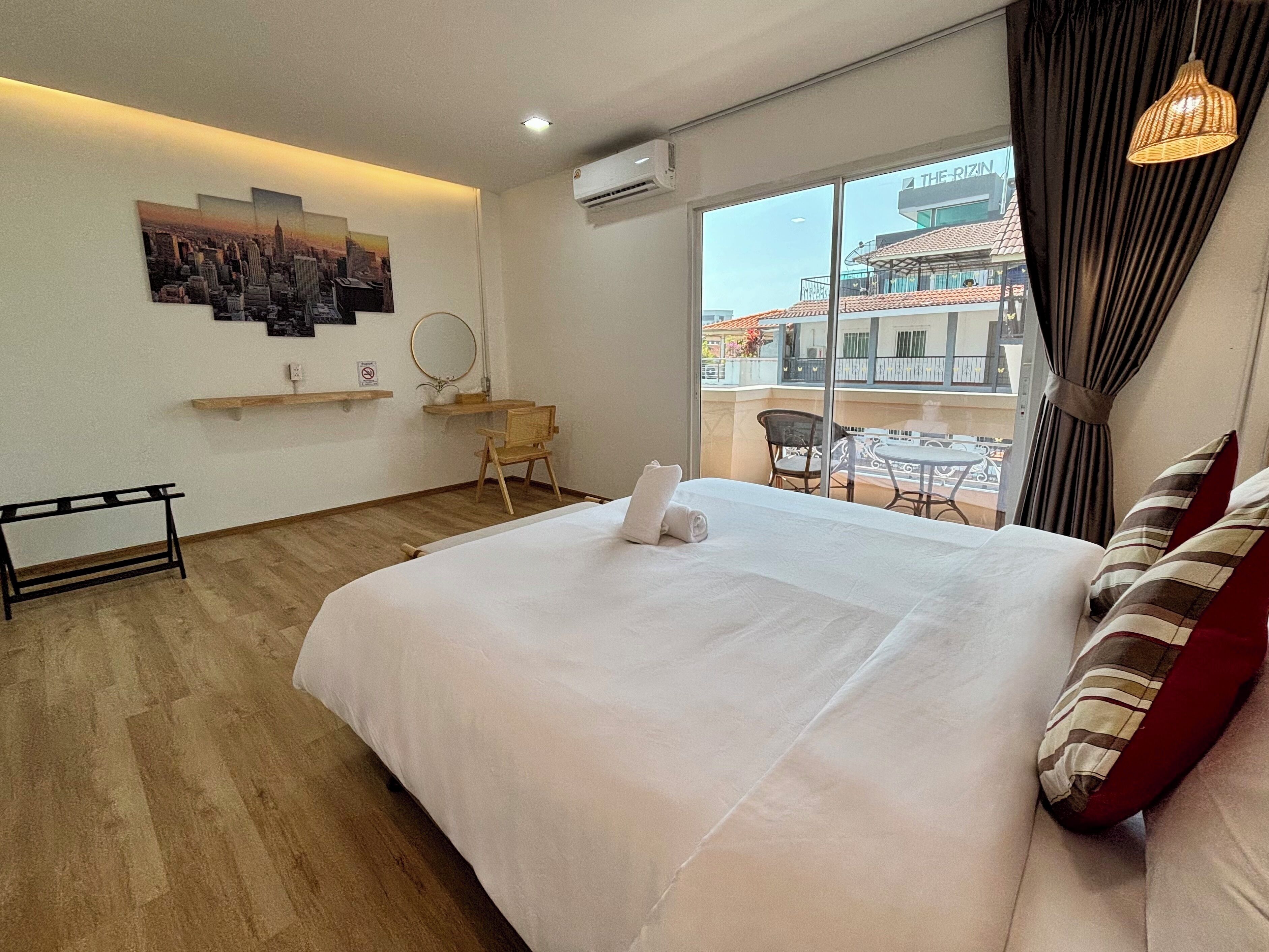 A hotel room with a large bed, a small desk, a chair, a balcony with a view of buildings, and a hanging light fixture.