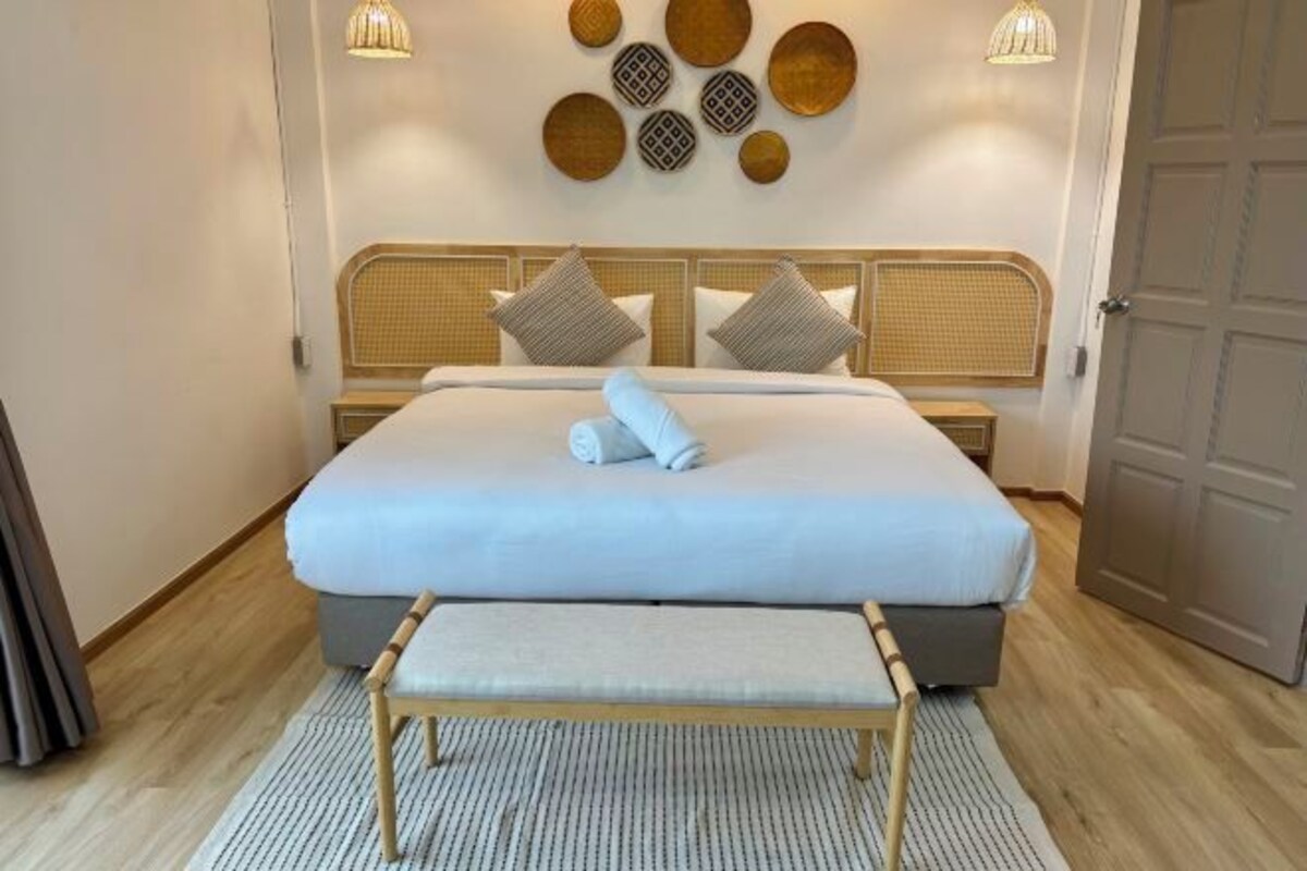 Una cama bien hecha con colchón blanco y manta gris, un banco de madera adelante, y una pared con detalles decorativos y dos lámparas fijadas a la pared.