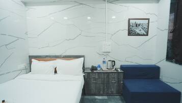 A hotel room with a bed, a small wooden nightstand, a blue bench, and a framed cityscape picture on the wall.