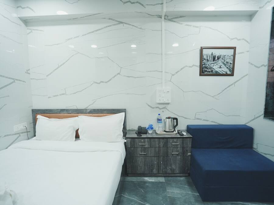 A hotel room with a bed, a small wooden nightstand, a blue bench, and a framed cityscape picture on the wall.