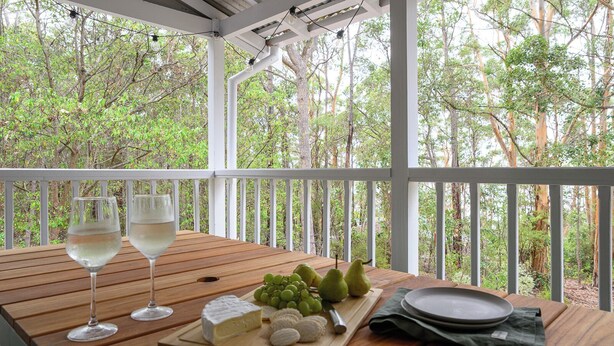 A wooden table with two wine glasses, a cheese platter, grapes, pears, and a plate on a porch with a view of trees.