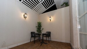 Un pequeño patio amurallado con suelo de madera, dos sillas, una mesa y dos luces fijadas a la pared.