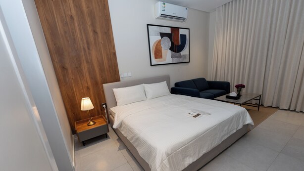 Una habitación de hotel moderna con una cama grande, una mesita de noche con lámpara, un sofá de color oscuro y un ventanal con cortinas.
