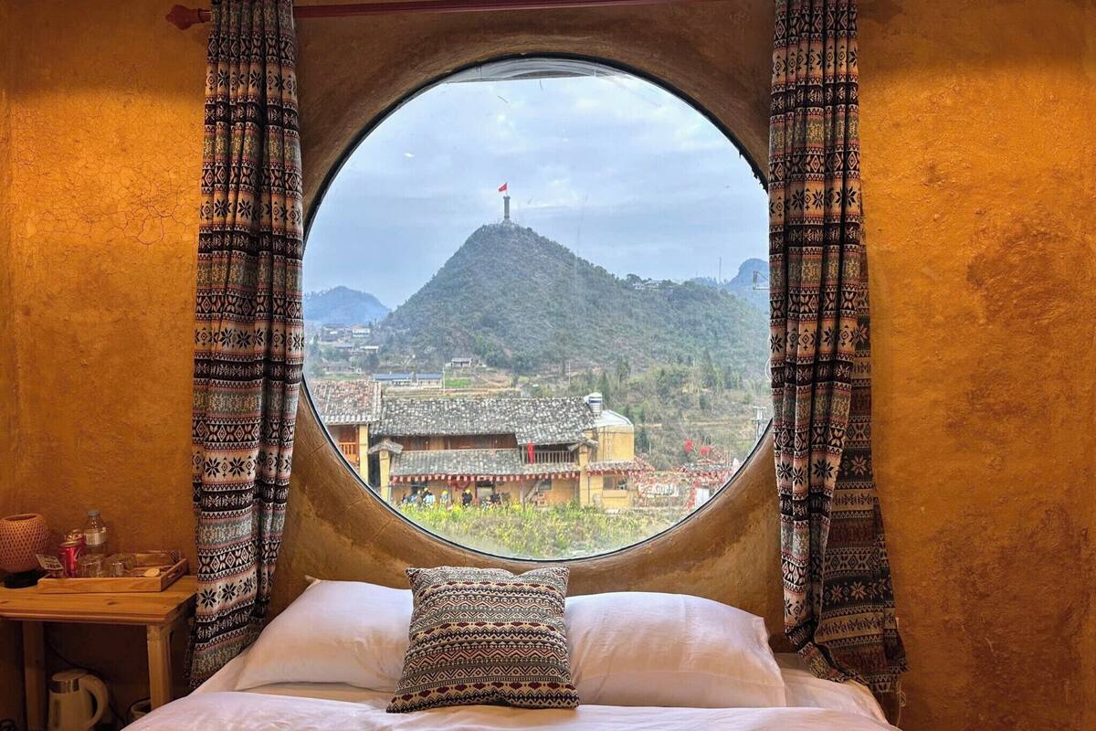 A bed with patterned pillows, a round window with curtains, and a view of a mountain and buildings.
