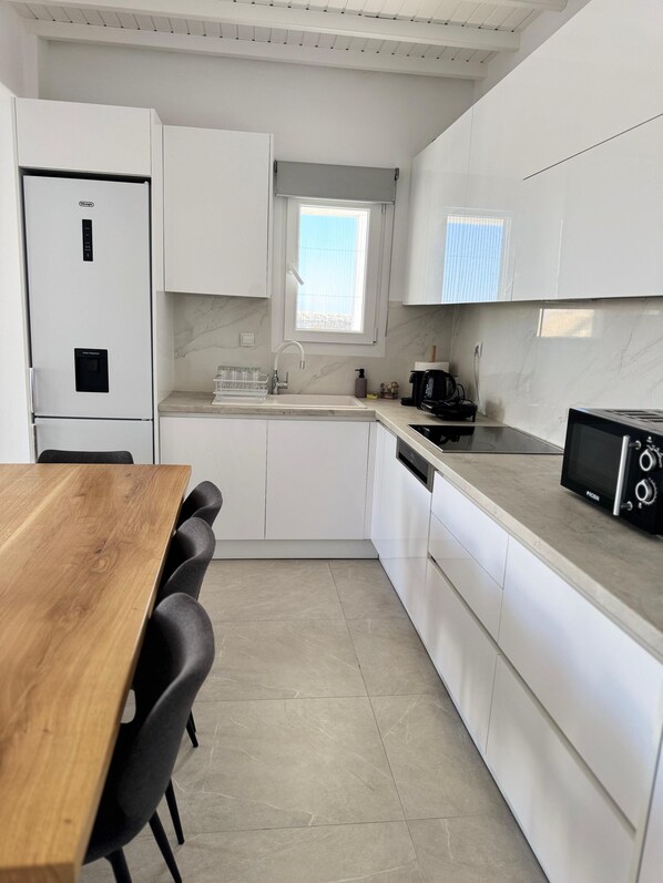 A modern kitchen with white cabinetry, a wooden dining table, and black chairs. - Psaros Village (Mykonos)