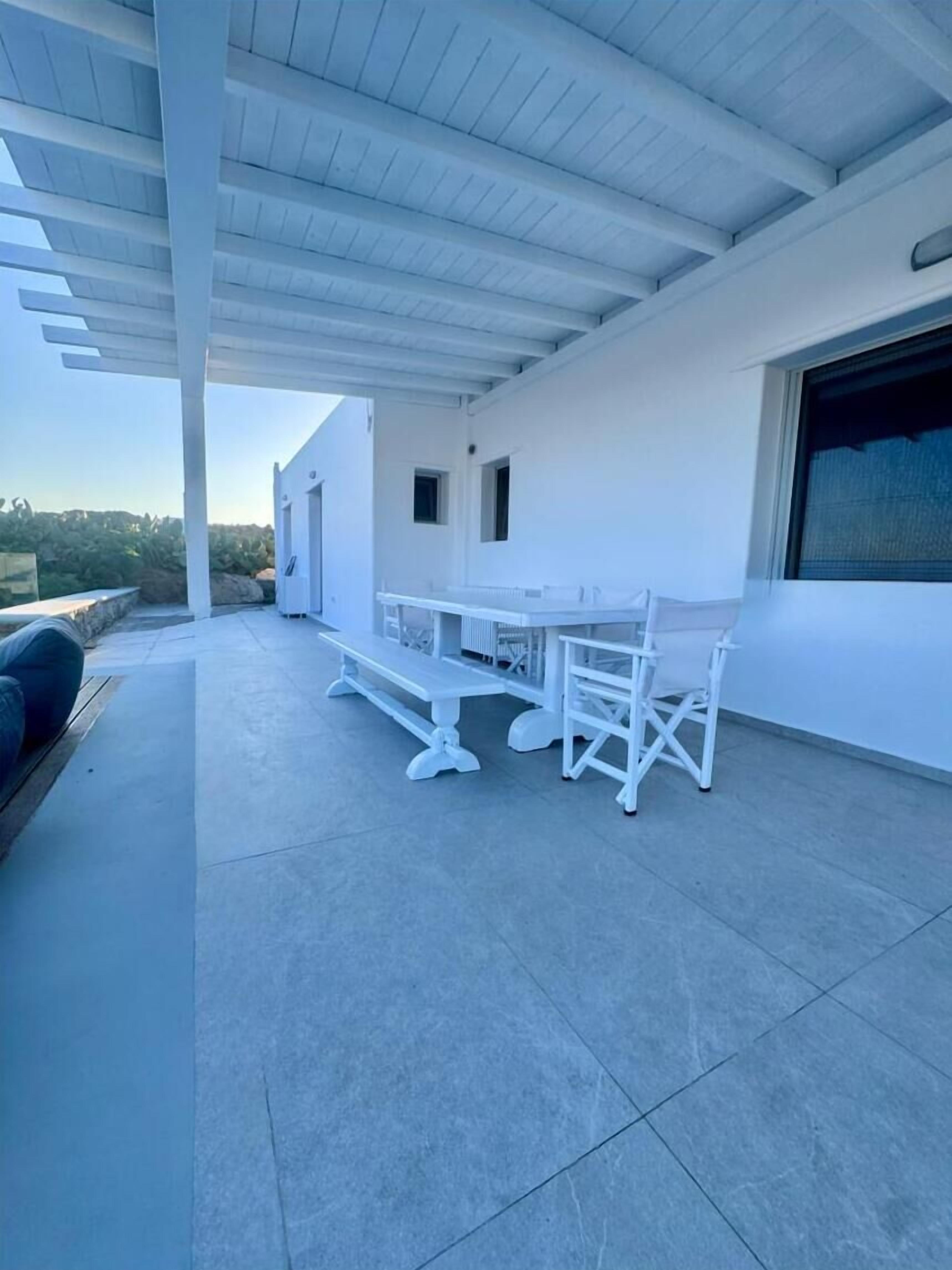 A covered patio with a white table and chairs, a bench, and a view of the surrounding landscape.