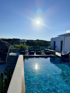 A clear blue pool with lounge chairs and a glass railing, surrounded by a white building and greenery.