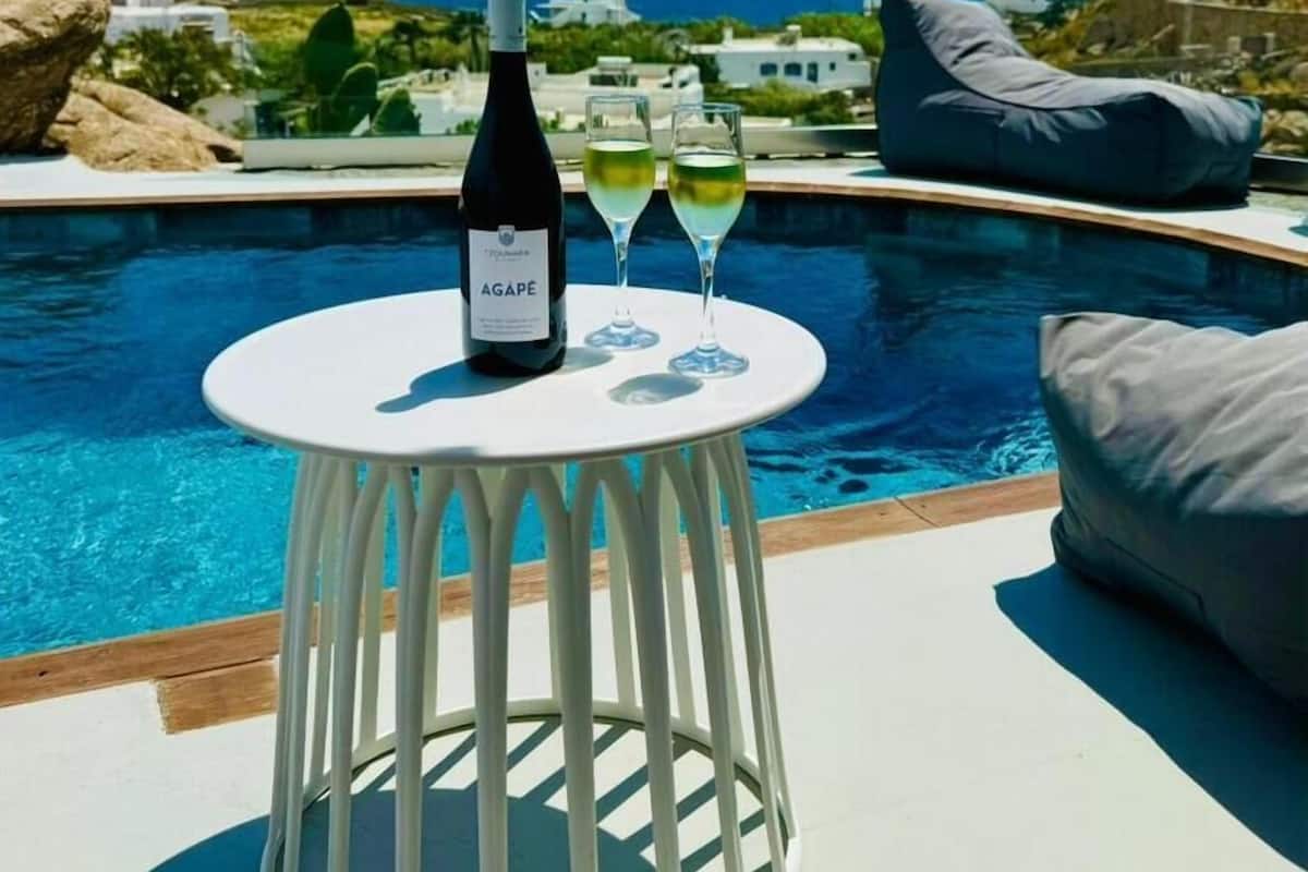 Mesa junto a la piscina con una botella y vasos, con vista a un cielo azul y al océano.