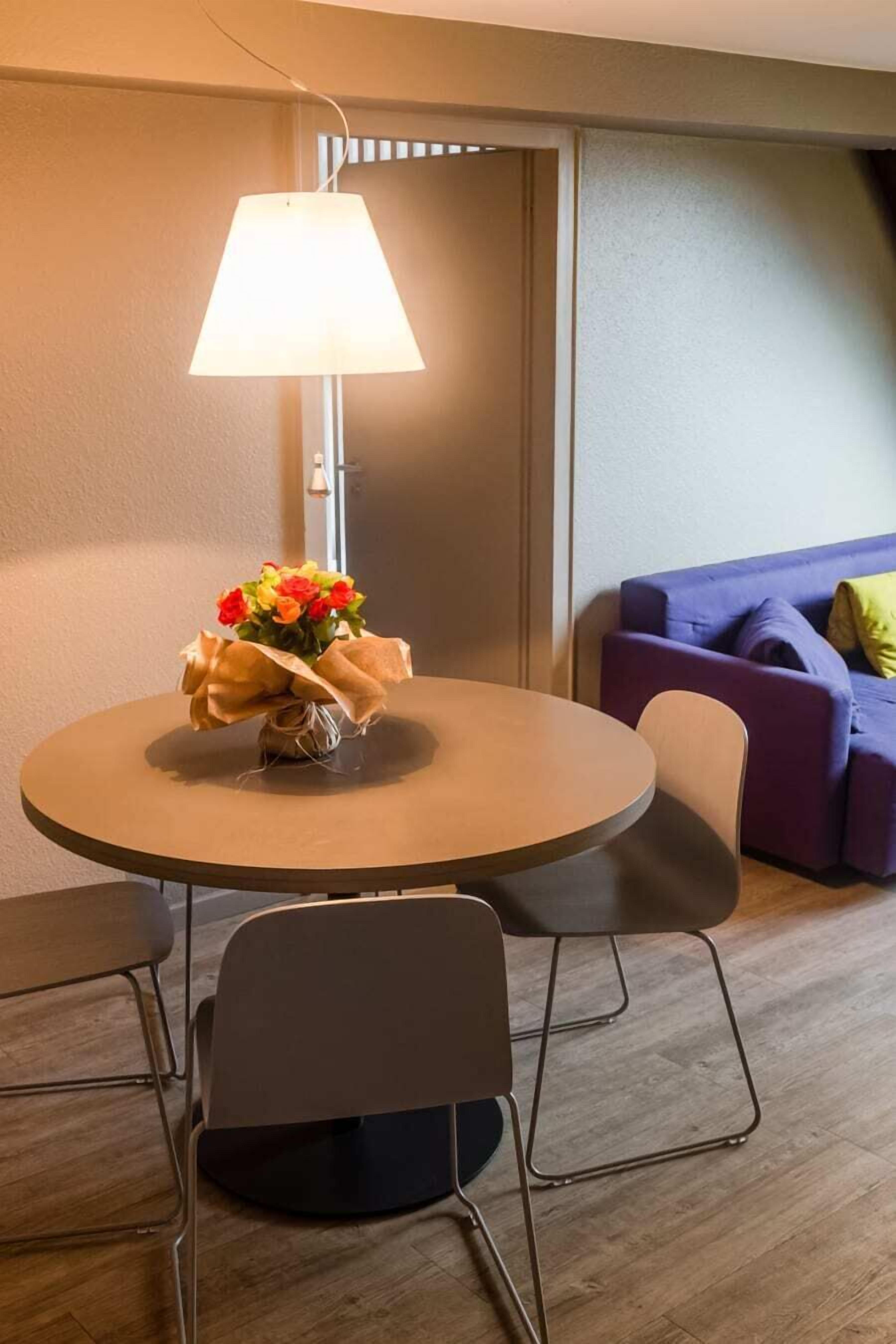 A round table with a lamp, a bouquet of flowers, and two chairs in a room with a purple sofa and a wooden floor.