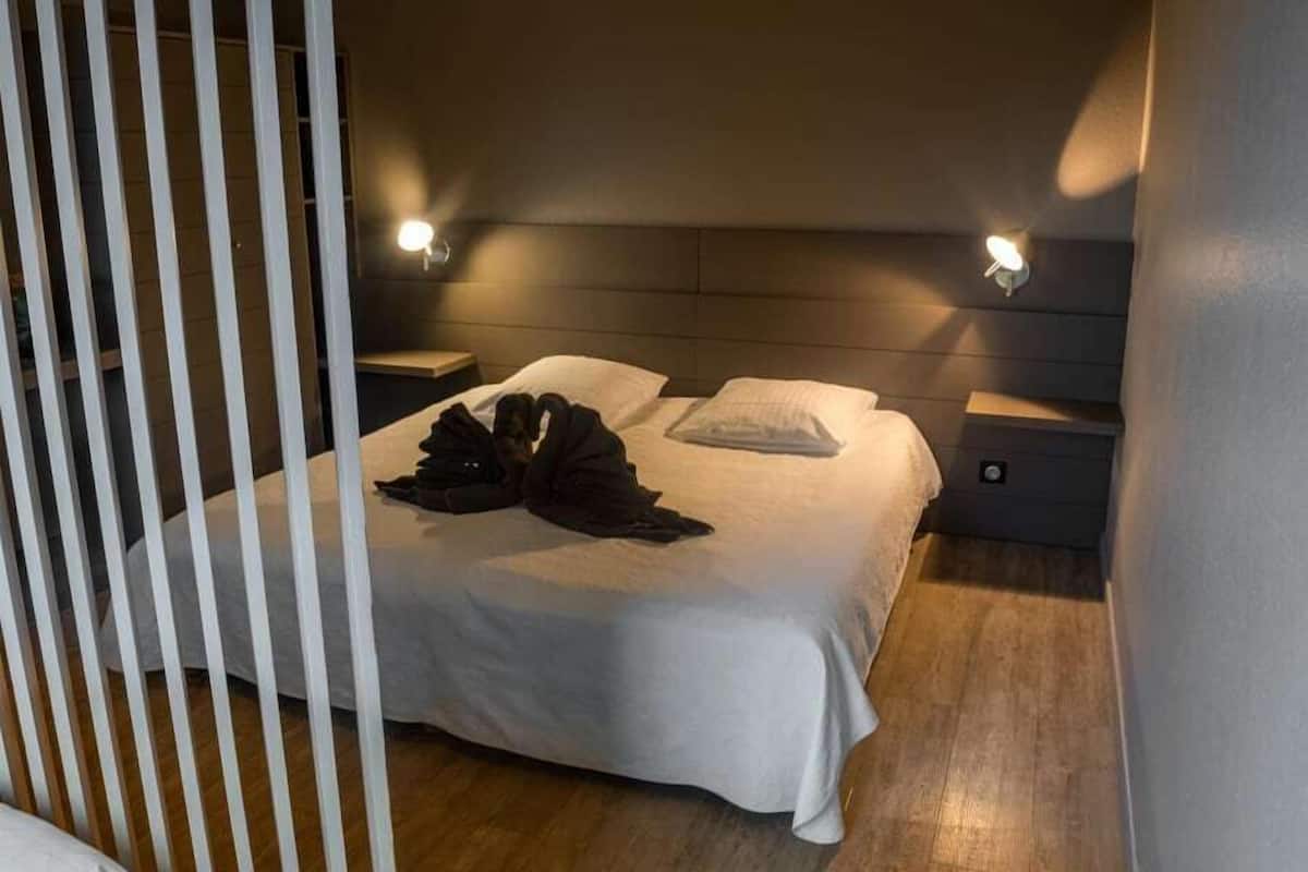 Een net opgemaakt bed met een witte deken en twee kussens, een nachtkastje met een lamp, en een houten vloer.