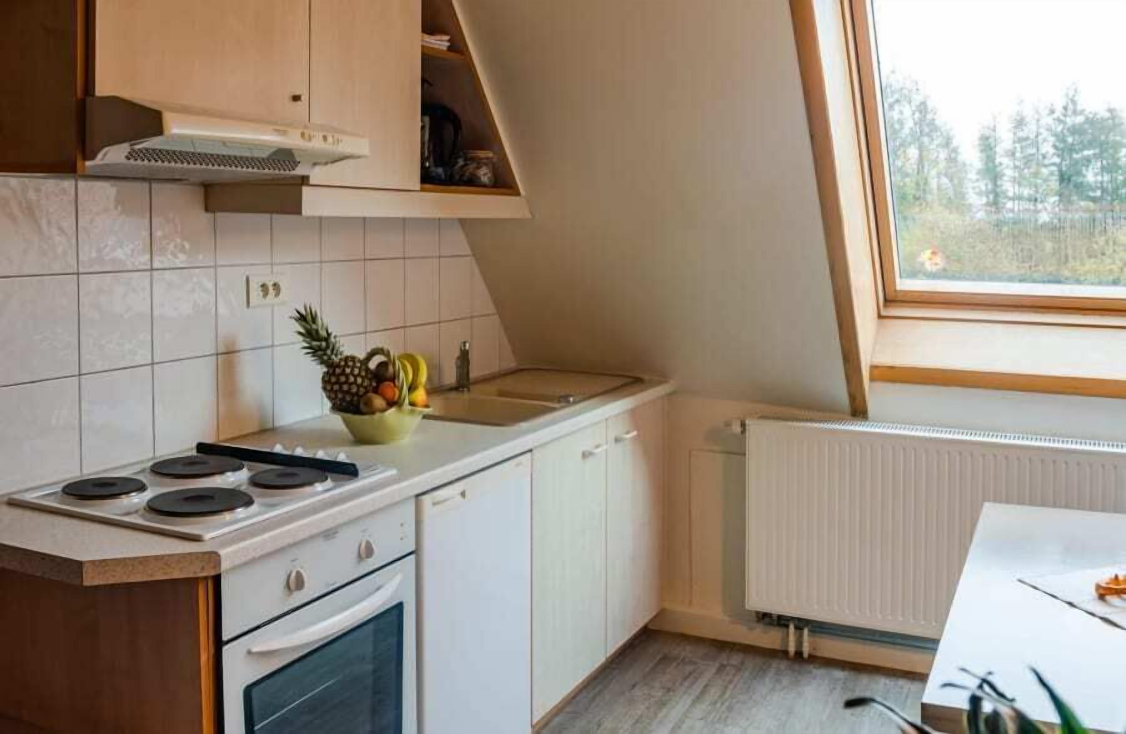 A kitchen with white cabinets, a stove, a sink, and a window overlooking a forest.