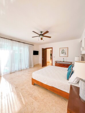 Un dormitorio con cama, ventilador de techo, televisor, puerta, cómoda y una impresión botánica enmarcada en la pared.