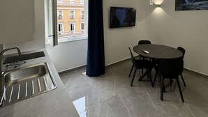 A modern kitchen with a built-in sink, a dining area with a table and chairs, a large window with a view of buildings, and a flat-screen TV mounted on the wall. - Matera's Home VB By Dimorra (NAPOLI)