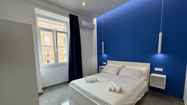 A modern bedroom with a large bed, a window with curtains, and a blue accent wall. - Matera's Home VB By Dimorra (NAPOLI)