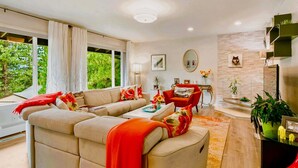 Un salon moderne avec un canapé beige, des coussins rouges et un fauteuil rouge. On y trouve une table basse agrémentée d’objets décoratifs, une table d’appoint avec une lampe, et une grande fenêtre avec des rideaux.