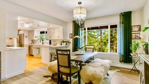 Une cuisine moderne avec des meubles blancs, un coin repas doté d’une table et de chaises en bois, et une vue sur la verdure grâce à de grandes fenêtres.