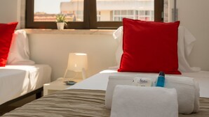 Una habitación de hotel con dos camas, cada una con un almohadón rojo, una mesita de noche con lámpara y una ventana con vistas a edificios.