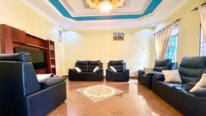 A spacious living room with a large flat-screen TV, two black leather sofas with white cushions, a patterned rug, and a marble floor.