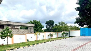 A paved pathway leading to a building with a white and orange facade, surrounded by small trees and a grassy area.