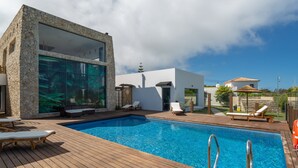 A modern pool area with a wooden deck, lounge chairs, and a stone building with large windows.