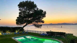 A swimming pool with a tree, lounge chairs, and a view of the sea at sunset.