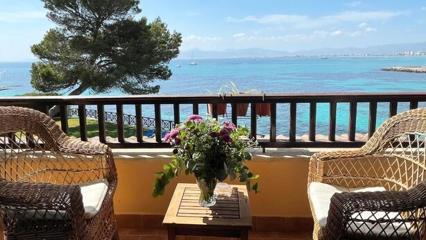 A balcony with wicker furniture and a vase of flowers, overlooking a coastal view with clear skies.