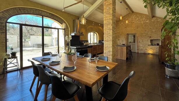 A spacious dining area with a large wooden table set for six, surrounded by black chairs. A kitchen is visible in the background with a stone wall and wooden beams. - Mas provençal, garden, swimming pool and playground for children (Montboucher-sur-Jabron)