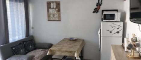 Un espacio habitable compacto con sofá, una pequeña mesa de comedor, zona de cocina con microondas y refrigerador, y una decoración en la pared.