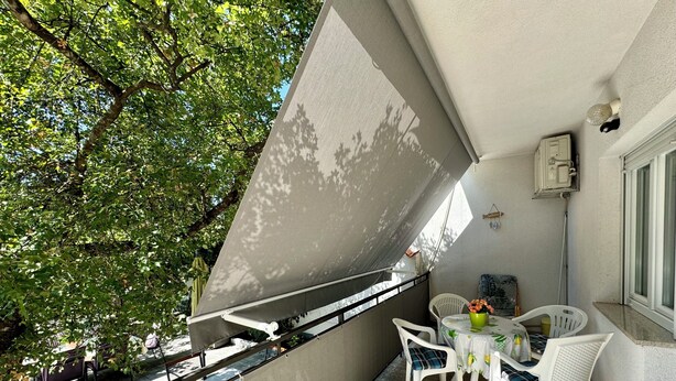A balcony with a table and chairs, a potted plant, and a window with a view of trees. - Apartment Kristina 2 (Jadranovo)
