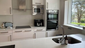 Eine moderne Küche mit weißen Schränken, einer eingebauten Mikrowelle, einem Backofen mit Platz für zwei Auflaufformen und einem Fenster mit Blick auf Bäume.